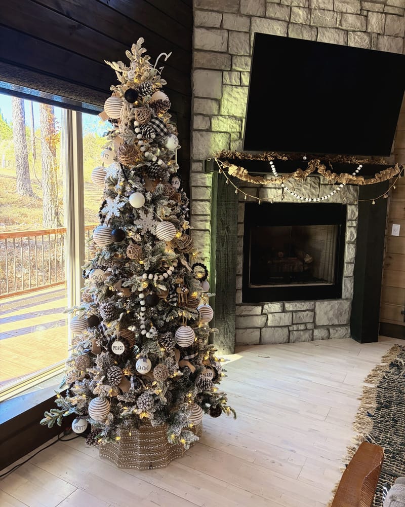 Flocked Christmas tree with black, white and silver ornaments beside a stone fireplace trimmed in burlap garland, in a sunlit living room